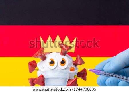 Funny cell of Covid-19 virus in medical mask near flag of Germany and a hand with syringe making an injection to it. Coronavirus vaccination in Germany