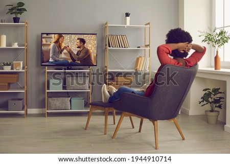 Woman relaxing in chair at home and enjoying television soap opera with a happy ending. Young lady sitting in comfortable armchair in living-room and watching romantic series or reality show on TV Royalty-Free Stock Photo #1944910714