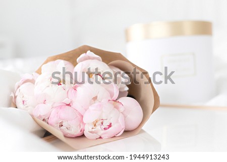 Gold gift box and white frame mockup on the bed. Bouquet of pink peonies in craft packaging. Scandivanavian white interior.