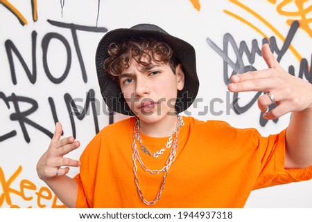 Yo yo look at me. Cool boy teenager with curly hair dressed in stylish clothes being street artist or rapper wears hat t shirt chains on neck poses against graffiti wall in urban area. Swag. Royalty-Free Stock Photo #1944937318