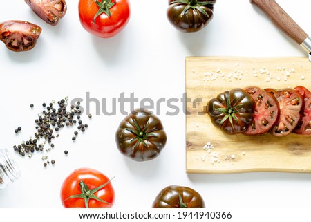 Cut and whole dark red and bright red tomatoes on wooden cutting board and black peppercorns on white background