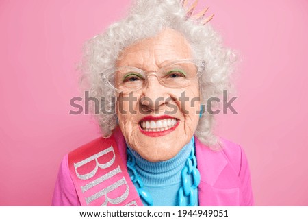 Close up shot of senior grey haired European woman smiles broadly applies bright makeup shows her perfect whit teeth celebrates birthday isolated over pink background. People old age concept