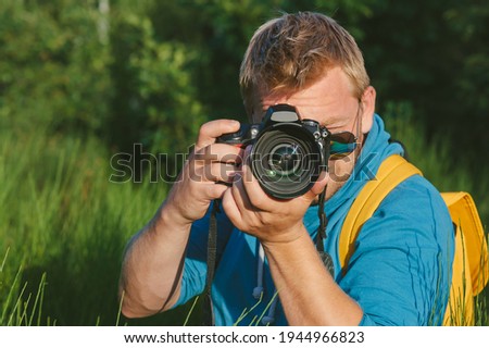 Close-up, the photographer holds a professional photo-video camera in his hands. Against the background of green beautiful nature