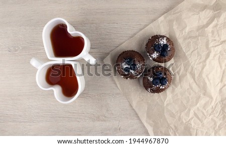 Three blueberry muffins and two heart shaped mugs with tea on wooden background. Tea party for two