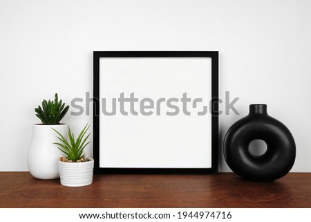 Mock up black square frame with succulent plants and modern circle vase. Wooden shelf against a white wall. Copy space.