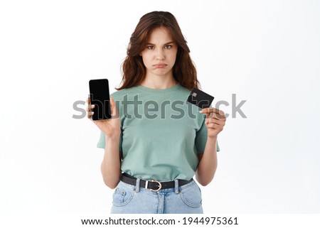 Sad and gloomy young girl showing smartphone empty screen and plastic credit card, empty bank account balance on mobile banking app, standing against white background