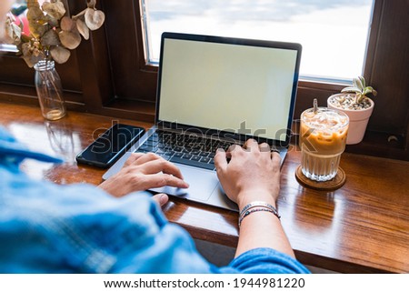 Close-up of male hands using laptop and smartphone at coffee shop with white mockup screen, man's hands typing on laptop keyboard, side view of a man using computer in cafe