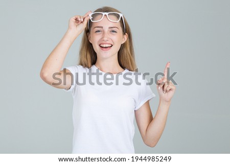 Young woman student raised her glasses pointing finger up and looks at the camera. Isolated on a gray background.