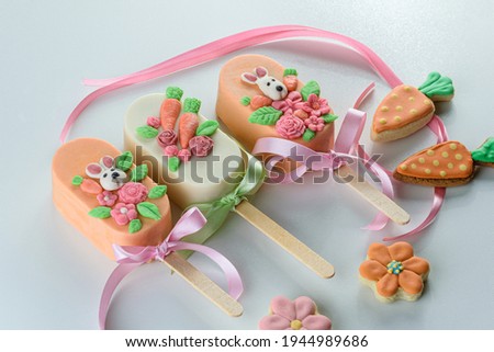 carrot cake cakesicles, decorated with rabbit, flowers and ribbon bow. Next to carrot and flower shaped cookies on a white background.