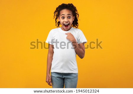 Who, Me. Portrait Of Shocked Surprised African American Girl Pointing Finger At Herself Posing Standing Isolated Over Yellow Orange Studio Background, Amazed Teen Smiling To Camera Royalty-Free Stock Photo #1945012324