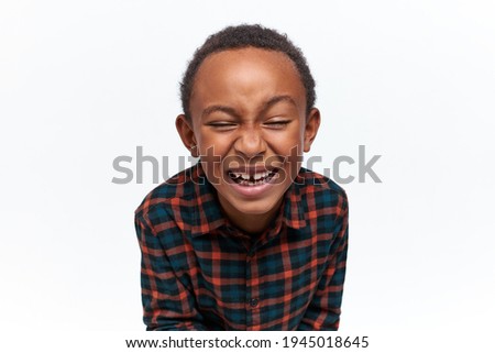Funny adorable African little boy wearing stylish plaid shirt having cheerful look, closing eyes, laughing out loud, watching favorite cartoon, showing milk teeth. Fun, joy and carefree childhood