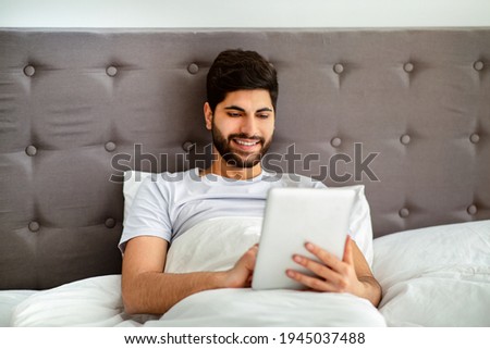 Weekend leisure concept. Relaxed arab man using digital tablet, sitting in bed and surfing internet online in bedroom at home. Gadget, technologies and modern lifestyle.