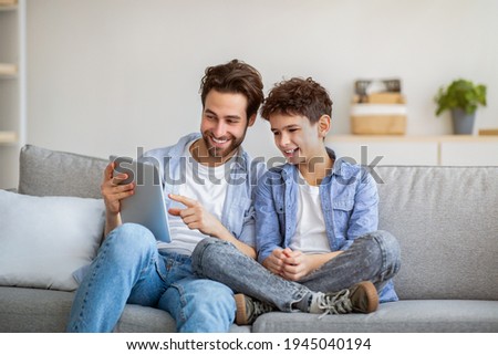 Joyful father and son using tablet computer, watching videos or playing online games, spending time together at home, sitting on comfy sofa. Leisure activities, modern technologies concept