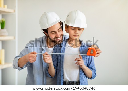 Construction working and repairs. Cheerful dad and his son using measuring tape and wearing safety helmets, dad teaching boy using roulette. Family spending time together at home Royalty-Free Stock Photo #1945040746