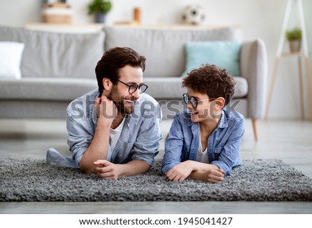 Men family. Cheerful father and son lying on floor carpet and lookig at each other, wearing eyeglasses and smiling, spending time together at home, enjoying weekend. Fatherhood concept Royalty-Free Stock Photo #1945041427