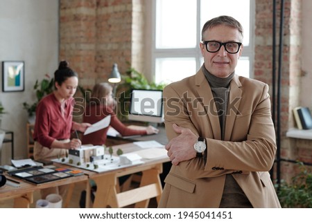 Smiling mature male architect in smart casualwear and eyeglasses standing against his two young colleagues working over model of new house