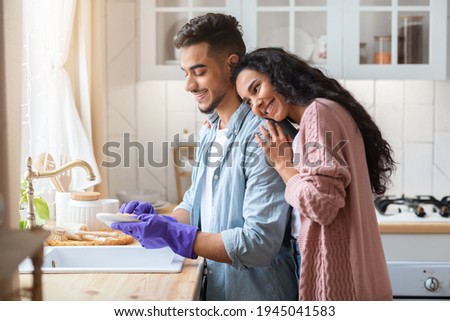 Young Arab Husband Washing Dishes After Lunch, Helping Wife With Domestic Chores. Middle Eastern Family Cleaning In Kitchen After Having Meal Together, Grateful Woman Hugging Husband, Free Space