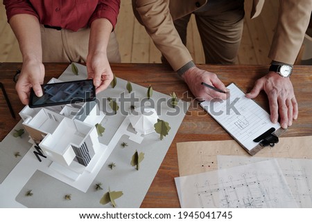 Hands of young female architect taking photo of model of new house and yard while her mature male colleague making notes below sketch on paper