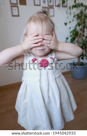small child in a white dress, baby, a blonde girl mischievously covered her face with her palms, plays hide and seek, the concept of childhood, parenthood, family everyday life