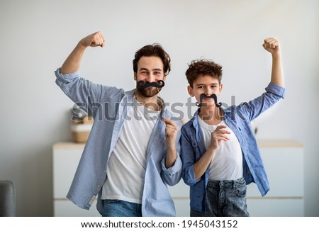Son and strong dad showing biceps, holding fake moustache on sticks and smiling to camera. Father and boy having fun, spending time together at home. Royalty-Free Stock Photo #1945043152