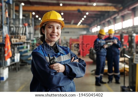 Young smiling female worker of modern industrial plant or factory in workwear and protective helmet standing in large workshop Royalty-Free Stock Photo #1945045402
