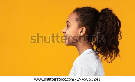 Side view profile portrait of cute African American girl with beaming smile and curly hair in ponytail looking aside at empty free copy space isolated on yellow studio background, banner, panorama Royalty-Free Stock Photo #1945053241