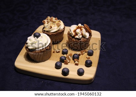 Three cupcakes with white buttercream on a wooden board among pecan nuts and blueberries
