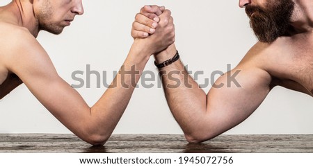 Two man's hands clasped arm wrestling, strong and weak, unequal match. Heavily muscled bearded man arm wrestling a puny weak man. Arms wrestling thin hand, big strong arm in studio.