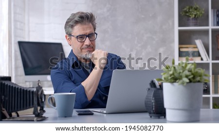 Bearded man working online with laptop computer at home sitting at desk. Businessman in home office, browsing internet. Portrait of mature age, middle age, mid adult man in 50s. Royalty-Free Stock Photo #1945082770