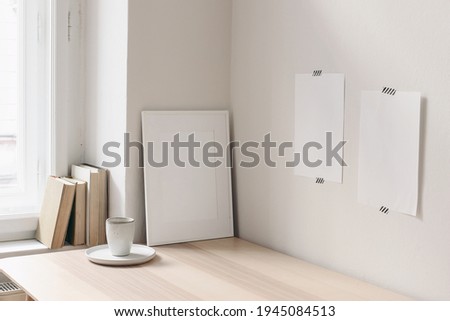 Breakfast still life. Cup of coffee, books on wooden desk, table near window. Empty notepads, posters taped on white wall.Blank picture frame mock-up. Elegant Scandinavian working space, home office. Royalty-Free Stock Photo #1945084513