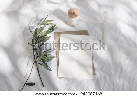 Summer wedding stationery, birthday mock-up scene. Blank greeting card with envelope, silk ribbon and olive tree branches. White linen textile background in sunlight, long shadows. Flat lay, top view.
