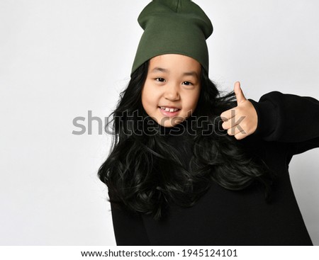 Young attractive preteen asian girl in warm autumn or spring black sportswear and dark green hat posing for camera showing thumbs up gesture approving expression. Kids emotion studio portrait