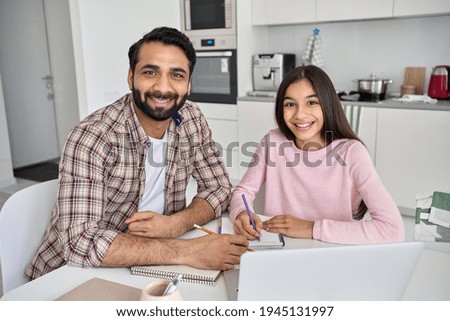 Happy young indian parent father helping teenage child daughter distance learning online together at home. Teen school kid girl studying in kitchen with dad looking at camera sitting at kitchen table.
