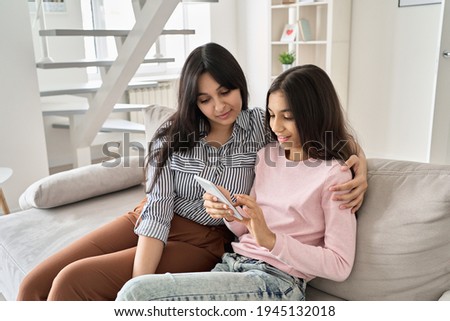 Happy indian family mom with teen daughter using phone together at home. Young mother and teenage child girl watching social media videos, doing online shopping on smartphone sitting on sofa together.