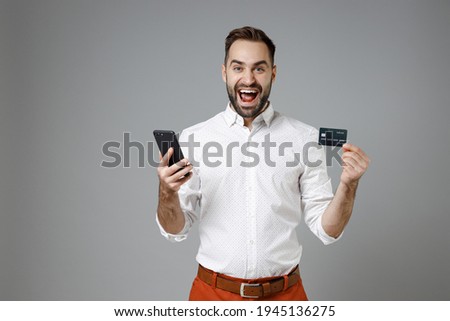 Excited young bearded business man in classic white shirt standing using mobile cell phone hold credit bank card isolated on grey color background studio portrait. Achievement career wealth concept