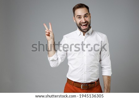 Excited laughing cheerful young bearded business man 20s wearing classic white shirt standing showing victory sign isolated on grey color background studio portrait. Achievement career wealth concept