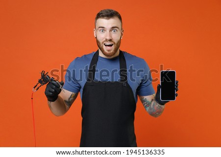 Amazed professional tattooer master artist man in t-shirt apron hold machine black ink in jar equipment for making tattoo art on body mobile phone with blank empty screen isolated on brown background