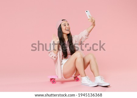 Full length portrait of funny young asian woman girl in cap isolated on pastel pink wall background. People lifestyle concept. Sit on skateboard, doing selfie shot on mobile phone, greeting with hand
