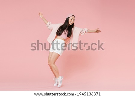 Full length portrait of funny young asian girl in casual clothes, cap isolated on pastel pink background studio. People lifestyle concept. Mock up copy space. Dancing spreading hands standing on toes