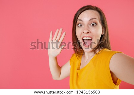 Close up of excited young brunette woman 20s in yellow casual t-shirt doing selfie shot on mobile phone waving greeting with hand keeping mouth open isolated on pink color background studio portrait