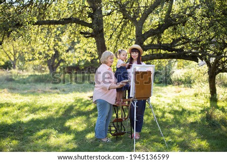 Beautiful summer spring shot of happy three generations family, little kid girl, young mother and mature grandmother, spending time outdoors and painting picture on easel. Summer family leisure.