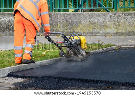 Paving worker uses vibratory plate compactor to compact new asphalt near curbstones  Royalty-Free Stock Photo #1945153570
