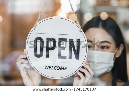 beautiful female business retail owner in medical face mask hanging open wooden sign board at the entrance door of the shop and ready to service customer.