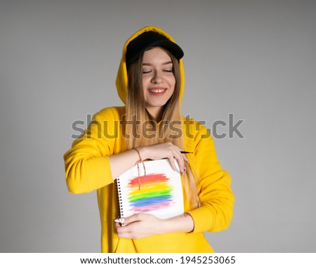 Happy girl in a yellow hoodie, a black cap with a pierced nose, holds a notebook on which an LGBTQ rainbow is drawn