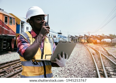 Portrait of handsome Africa American engineering using walkie talkie and laptop for control labor in front of train garage.  Back view of contractor on background of outdoor old train garage. Royalty-Free Stock Photo #1945253767