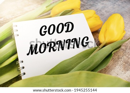 Good morning is written on a notebook that lies among a bunch of tulips on a wooden table.