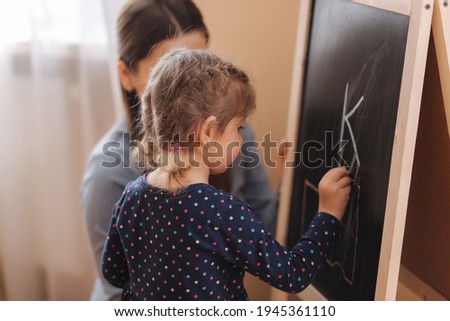 Cute little daughter with her young mom learn writes a letters on the blackboard at home. Side view of female kid in dress learning