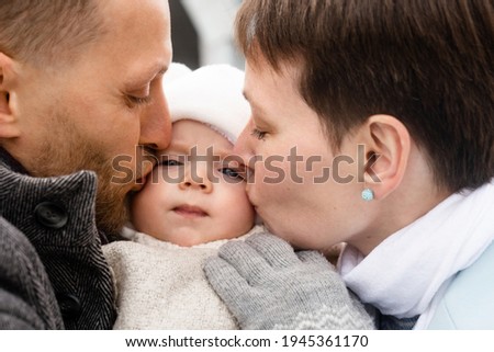Tender kiss. Portrait view of the brunette short haired woman and handsome unshaven man holding their little son at the hands and kissing him at the chick. Happy parenthood concept. Stock photo