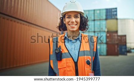 Smiling Portrait of a Beautiful Hispanic Female Industrial Engineer in White Hard Hat, Safety Vest and with Two-Way Radio Working in Logistics Center. Inspector or Supervisor in Container Terminal. Royalty-Free Stock Photo #1945399777