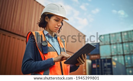 Smiling Portrait of a Beautiful Latin Female Industrial Engineer in White Hard Hat, High-Visibility Vest Working on Tablet Computer. Inspector or Safety Supervisor in Container Terminal. Royalty-Free Stock Photo #1945399801